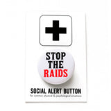 A round white pinback button that reads STOP THE RAIDS in black & red text. Button is on a Social Alert Button backing card.