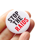 Round white pinback button that reads: STOP THE RAIDS in black & red text. Badge is held in a hand.