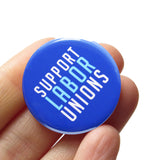 Dark blue round pinback button that reads Support Labor Unions in white & light blue text. Button is held in a hand.