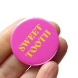Round vibrant pink pinback button reading SWEET TOOTH in yellow text. Pinned to a Social Alert Button backing card. Button held in a hand.