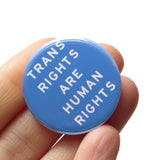 A round light blue button that read TRANS RIGHTS ARE HUMAN RIGHTS in white text. Button is held in a hand.