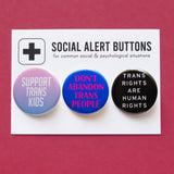 A Social Alert Buttons backing card with 3 pinback buttons attached, reading: Support Trans Kids, Don't Abandon Trans People, Trans Rights are Human Rights, in various shades of pink, blue and black.