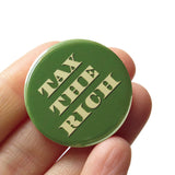 Round pinback button that says TAX THE RICH.  The text is a light green, on a money green background. Button is held in a hand.