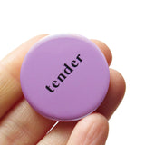 A round lavender 1.25" pinback button that reads tender in small, lower case black text. Button is held in a hand.