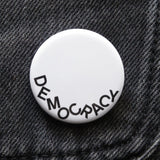 A round white pinback button that reads DEOMCRACY in black letters fallen to the bottom of the button.  Pinned on black denim.
