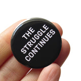 Round black pinback button that reads OUR FIGHT HAS JUST BEGUN in white text. Button is oheld in a white hand

