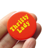 Round orange pinback button that reads THRIFTY LADy in a retro 70's yellow font. Button is held in a hand.