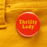 Round orange pinback button that reads THRIFTY LADy in a retro 70's yellow font. Button is pinned to yellow pants.