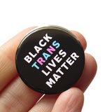 Round pinback button that says BLACK TRANS LIVES MATTER. White, light blue and light pink text on a black background. Button is held in a hand.