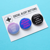 A Social Alert Buttons backing card with 3 pinback buttons attached, reading: Support Trans Kids, Don't Abandon Trans People, Trans Rights are Human Rights, in various shades of pink, blue and black.