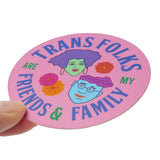 A round pink sticker that reads TRANS FOLKS ARE MY FRIENDS & FAMILY around the permitter of the sticker in periwinkle & green text, with two illustrated faces in the middle, one with minty skin, purple hair and pink lips. The other with blue skin, glasses and a wavy swoosh of magenta hair. Little pink, orange & yellow flowers are decorating the negative space around them. Sticker is held at an angle in the corner with a thumb and forefinger.