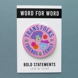 Large light-pink round pinback button, reads TRANS FOLKS ARE MY FRIENDS & FAMILY in blue & green text around the edge of button. Center has illustrated drawings of 2 heads with little flowers in the negative space. Head on the left is feminine with mint green skin, voluminous lavender hair, hoop earrings & pink lipstick. The face on the right is masculine with square jaw, light blue skin, round purple glasses, wavy pink hair, chin dimple & small smile. The button is on a Word For Word backing backing card