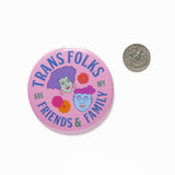 Large light-pink round pinback button, reads TRANS FOLKS ARE MY FRIENDS & FAMILY in blue & green text around the edge of button. Center has illustrated drawings of 2 heads with little flowers in the negative space. Head on the left is feminine with mint green skin, voluminous lavender hair, hoop earrings & pink lipstick. The face on the right is masculine with square jaw, light blue skin, round purple glasses, wavy pink hair, chin dimple & small smile. The button is next to a quarter for size comparison.