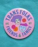 Large light-pink round pinback button, reads TRANS FOLKS ARE MY FRIENDS & FAMILY in blue & green text around the edge of button. Center has illustrated drawings of 2 heads with little flowers in the negative space. Head on the left is feminine with mint green skin, voluminous lavender hair, hoop earrings & pink lipstick. The face on the right is masculine with square jaw, light blue skin, round purple glasses, wavy pink hair, chin dimple & small smile. The button is on a a turquoise sweatshirt