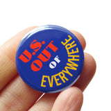 A round deep blue pinback button that reads US OUT (in red) OF (in white) and EVERYWHERE in yellow text curved around the bottom of the button. Badge is held in a hand.