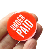 Bright round red-orange pinback button that reads UNDER PAID in white san serif text. Button held in a hand.