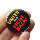 A round black pinback button that reads UNITE AND FIGHT BACK! in yellow, blue and red type. Button is held in a hand.