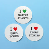 Three round white stickers on a light blue background. Top Sticker reads: I LOVE NATIVE PLANTS with love being represented by a green heart. Beneath reads I LOVE USED BOOKS with love being represented by a red heart. To the right reads I LOVE SHORT STORIES with love being represented by a red heart.
