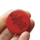 A round rust orange-red pinback button that reads SWE MUST PROTECT EACH OTHER in a thin black font. Button is held in a hand.