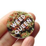 Round pinback button that reads WEED QUEEN in white text over a background of cannabis flower. Button is held in a hand.