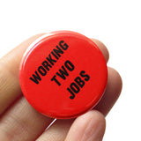 A round red-orange pinback button that reads WORKING TWO JOBS in black text. Button is held in a hand.