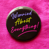 A round black  pinback button that reads WORRIED ABOUT EVERYTHING!  in purple, neon green & magneta pink text, in a retro 80's vibe.  Button is pinned to bold pink linen fabric.