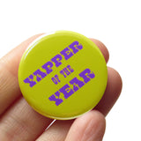 A round lime green pinback button that reads YAPPER OF THE YEAR in a chunky purple text. Held in a hand.