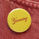 A round butter yellow 1.25" pinback button that reads " Yearning" in a Burgundy script font. The button is pinned to orange rust colored denim.
