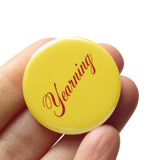 A round butter yellow 1.25" pinback button that reads " Yearning" in a Burgundy script font. The button is held in a hand.