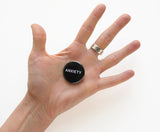 Round black pinback button that reads ANXIETY in white text.  Held in the palm of a hand with all 5 fingers outstretched in an anxious way