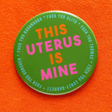 Big 3 inch shiny olive green pinback button with bright orange and hot pink chunky san serif text that reads: This Uterus is Mine. Smaller text in a light blue runs in a circle around the permitter that says Fuck You Alito, Fuck You Thomas, Fuck you Coney-Barrett, Fuck you Gorsuch, Fuck you Kavanaugh. The button is pinned to a bright orange sweater shirt. 