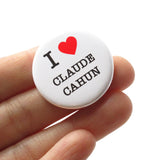 A round white pinback button that reads I LOVE CLAUDE CAHUN with love being a red heart. Held in a hand.