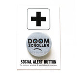 A round pinback button with a pale blue background of faded layered text, with thick black letters that say DOOM SCROLLER, the O's have half lidded eyes, and there's a subtle frown line drown underneath. Button is on a white backing card that reads Social Alert Button on the bottom, with a large plus sign at the top.