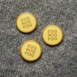 3 round buttery pale yellow enamel pins with gold colored text that reads EGG NOG FOG in three lines, and a clean gold circular outline. Pinned to a cozy gray wool sweater.