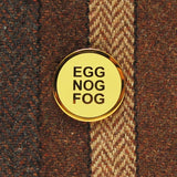 A round buttery pale yellow enamel pin with gold colored text that reads EGG NOG FOG in three lines, and a clean gold circular outline. Pinned to a stripped wool blazer with various shades of brown stripes.