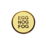 A round buttery pale yellow enamel pin with gold colored text that reads EGG NOG FOG in three lines, and a clean gold circular outline. Pin is on a white background.