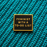 Gold & black rectangle enamel pin that reads FEMINIST WITH A TO-DO LIST. Pinned to a teal blue knitted sweater.