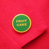 small circular enamel pin, green enamel background with gold text that reads FRUIT CAKE across two lines. Gold colored outline. Pin is on a red blazer lapel.