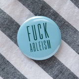 A round pale teal pinback button that reads FUCK ABLEISM in a dark teal color, in a condensed sans serif font.  Button is pinned to a gray & white diagonal stripped cotton shirt.