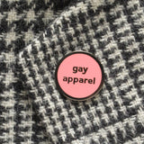 Small circular pink enamel pin with gunmetal silver text and outline that reads GAY APPAREL. Pin is pinned on a black and white checked coat.