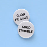 2 round off white pinback buttons that reads GOOD TROUBLE n black text. Buttons are on a light blue background, overlapping each other.