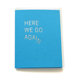 A bright medium blue greeting card with off white envelope, reads HERE WE GO AGAIN in a thin metallic silver foil, sans serif font. The word AGAIN starts to slide off, creating the image of the word coming apart and falling downward
