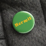 Shiny round dark green button with goldenrod serif text that reads: HERMIT. Hermit button is pinned to a puffy gray quilted  robe