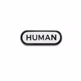 Capsule shaped hard enamel lapel pin with dark gray metal spelling HUMAN in a san serif font, with a dark metal outline. The pin is against a white background.