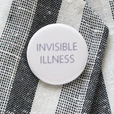 Round pinback button that reads INVISIBLE ILLNESS. Thin blue text on a white background. Button is pinned on white & gray woven blazer lapel.