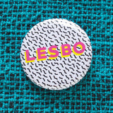 A round 1.25" white button with tiny Memphis style squiggles as a background pattern. Reading LESBO in bold pink & yellow text. Badge is pinned to a teal woven shirt.