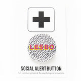 A round 1.25" white button with tiny Memphis style squiggles as a background pattern. Reading LESBO in bold pink & yellow text. Badge ispinned to a Social Alert Button backing card.