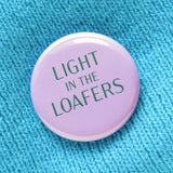 A round lavender pinback button that reads LIGHT IN THE LOAFERS in forest green. Button is on a aqua knit sweater.