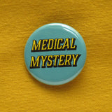 Round pinback button that says MEDICAL MYSTERY. Yellow shadowed text on a scrubs blue background. Button is pinned to a mustard yellow fabric.