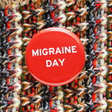 Shiny bright red pinback button with white san serif test reading, Migraine Day.  The button is pinned to a multicolor knit sweater with flecks of cream, black, brown read and pale blue.